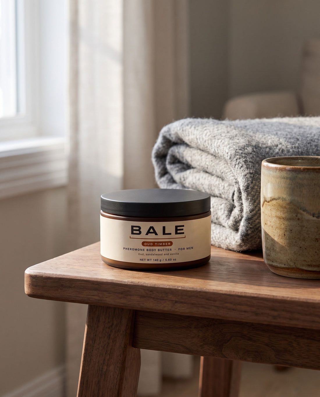 A jar of Bale Oud Timber body butter on a walnut table beside a wool blanket and a mug