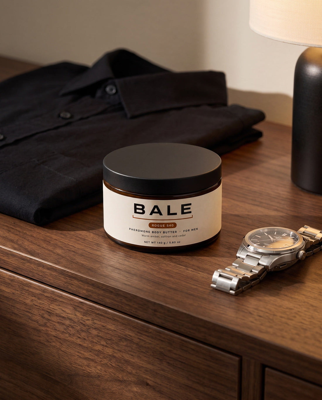 A jar of Bale Rogue 540 body butter on a dark wood dresser beside a black shirt and a watch