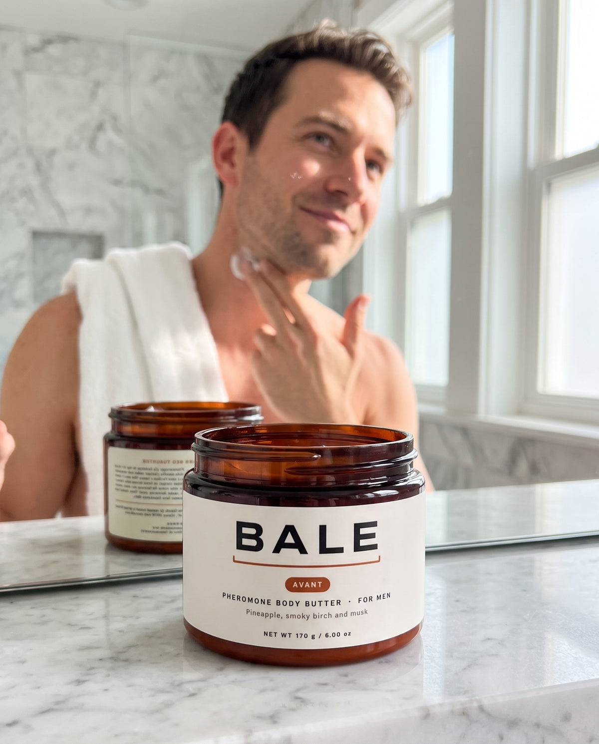 Man pressing Avant body butter into his neck in a bright bathroom, open jar on the counter