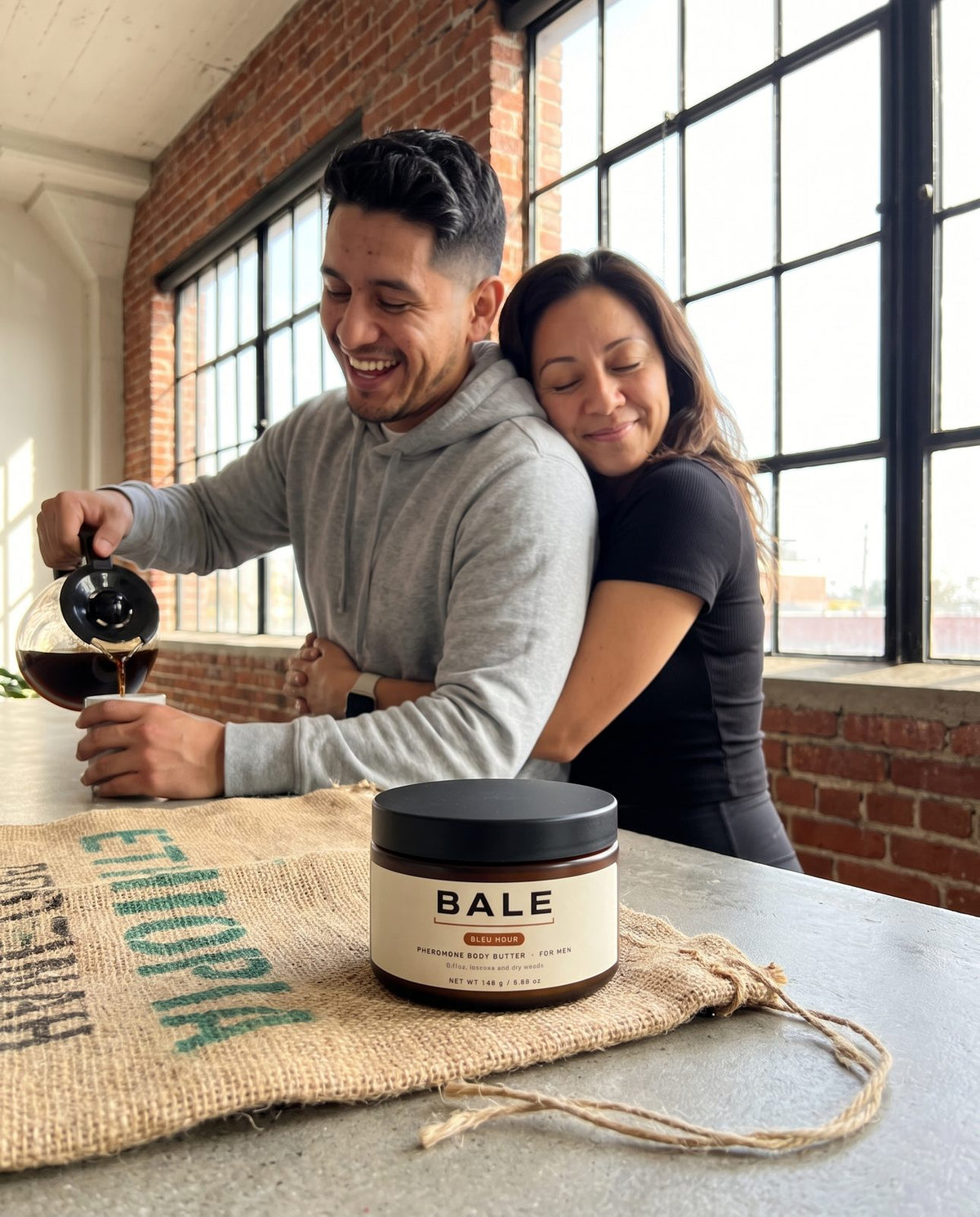 She hugs him from behind in a sunny kitchen, Bleu Hour jar on the counter
