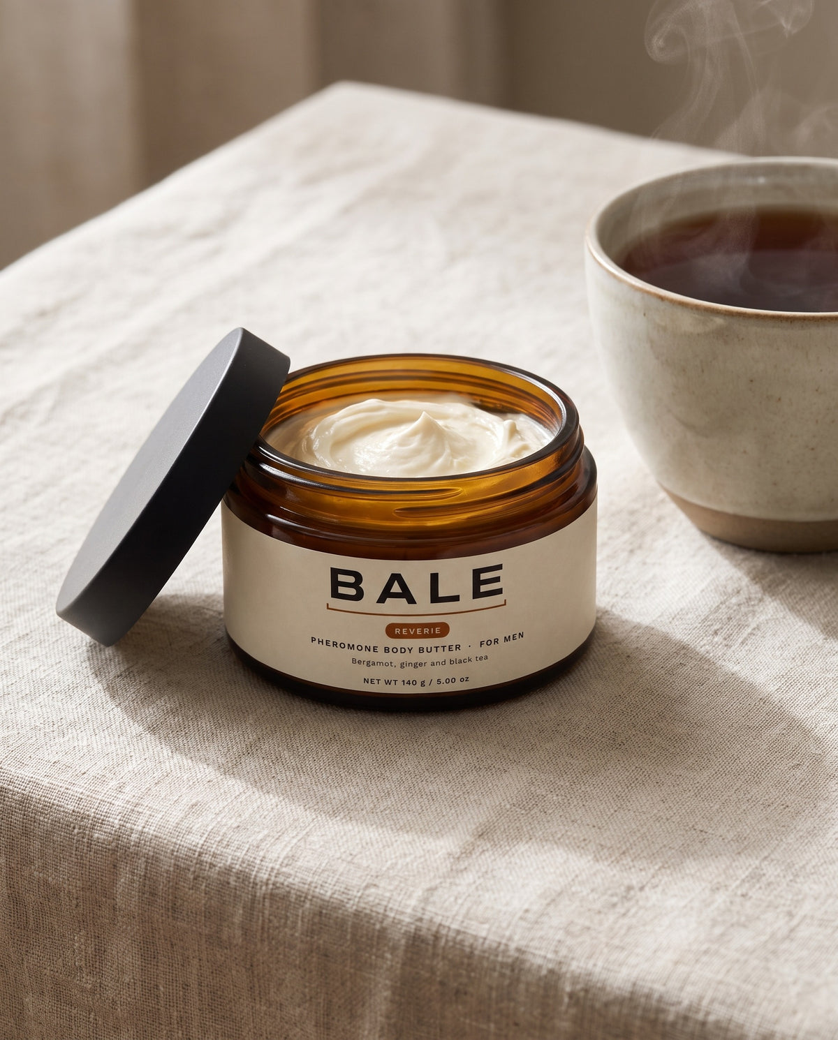 An open jar of Bale Reverie body butter on linen beside a cup of black tea