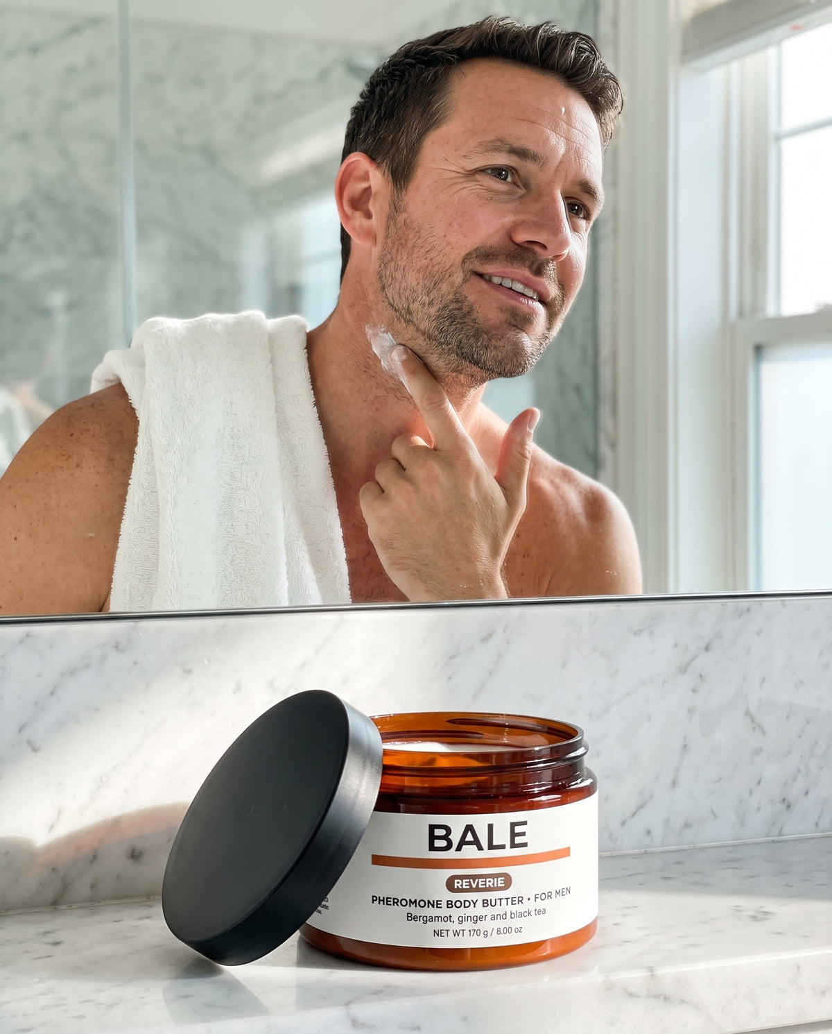 Man pressing Reverie body butter into his neck in a bright bathroom, open jar on the counter